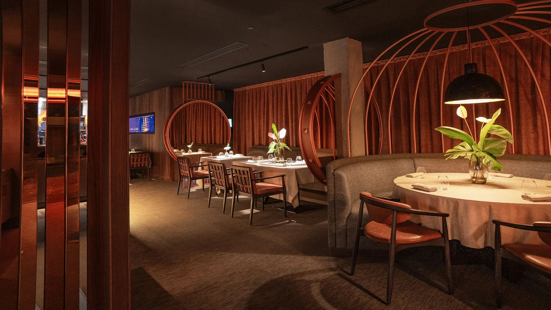 Comedor en penumbra con un reservado circular bajo una estructura de arcos de cobre, cortinas terracota y mesas con mantel blanco